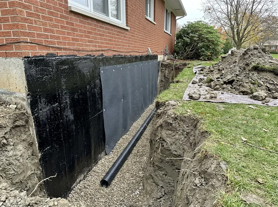 Excavated trench beside a basement wall with a fresh black waterproofing membrane, dimple board and new drain pipe at the footing